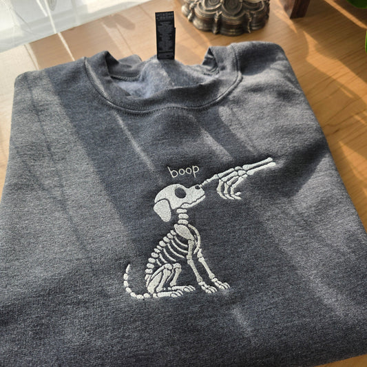A folded dark gray crewneck sweatshirt with a white embroidered dog skeleton graphic and the word 'boop' above it, placed on a wooden table.