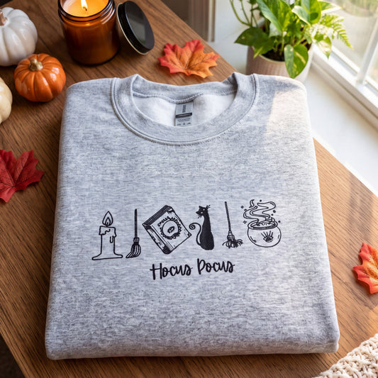A folded heather gray crewneck sweatshirt with a black 'Hocus Focus' graphic featuring Halloween-themed icons, displayed on a wooden table with autumn props like pumpkins, leaves, and a candle.