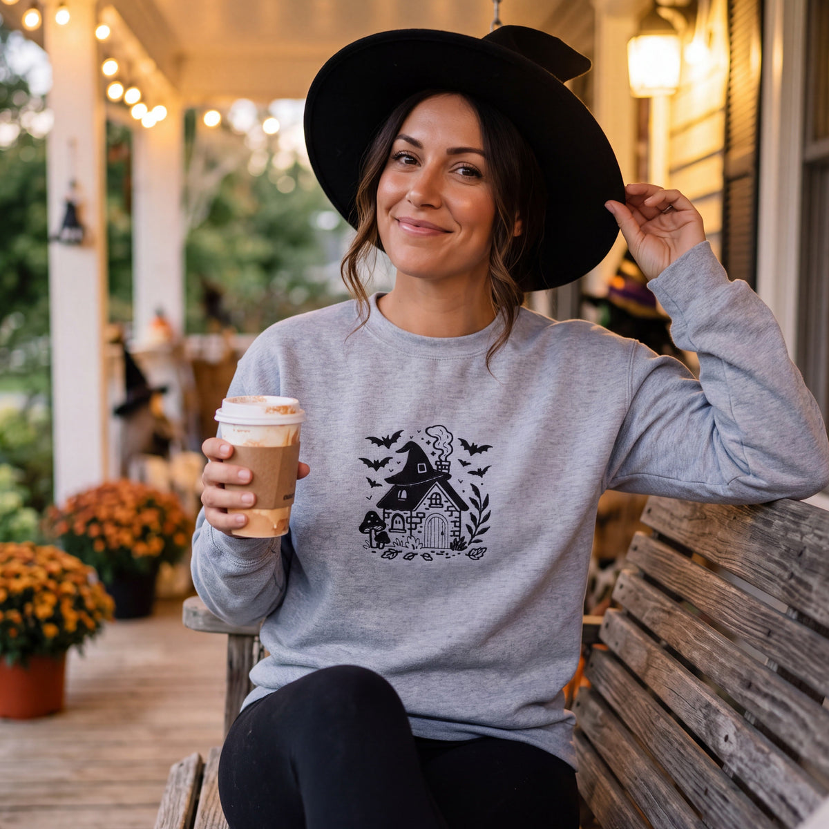 A woman sitting on a wooden porch swing wearing a light gray crewneck sweatshirt with a black witch graphic, a black witch hat, and holding a takeaway coffee cup.