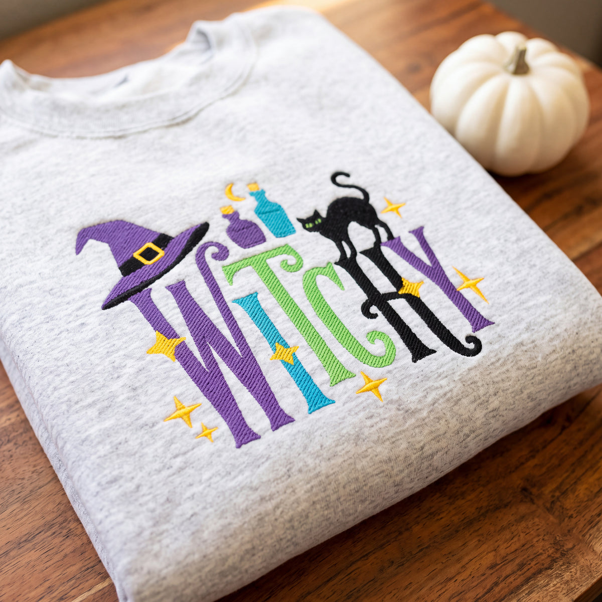A folded light heather gray t-shirt with a colorful embroidered 'Witch' graphic, displayed on a wooden table next to a small white pumpkin.