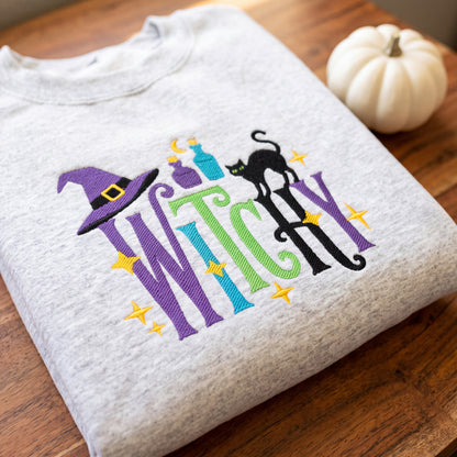 A folded light heather gray t-shirt with a colorful embroidered 'Witch' graphic, displayed on a wooden table next to a small white pumpkin.