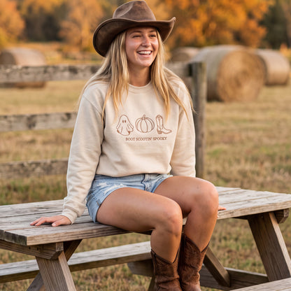 A woman sitting on a wooden picnic table outdoors in a fall field, wearing a cream sweatshirt with a brown graphic print that reads 'ROOT SCOUTIN' SPOOKY' with ghost, pumpkin, and cowboy boot illustrations, along with denim shorts, a brown cowboy hat, and brown cowboy boots.